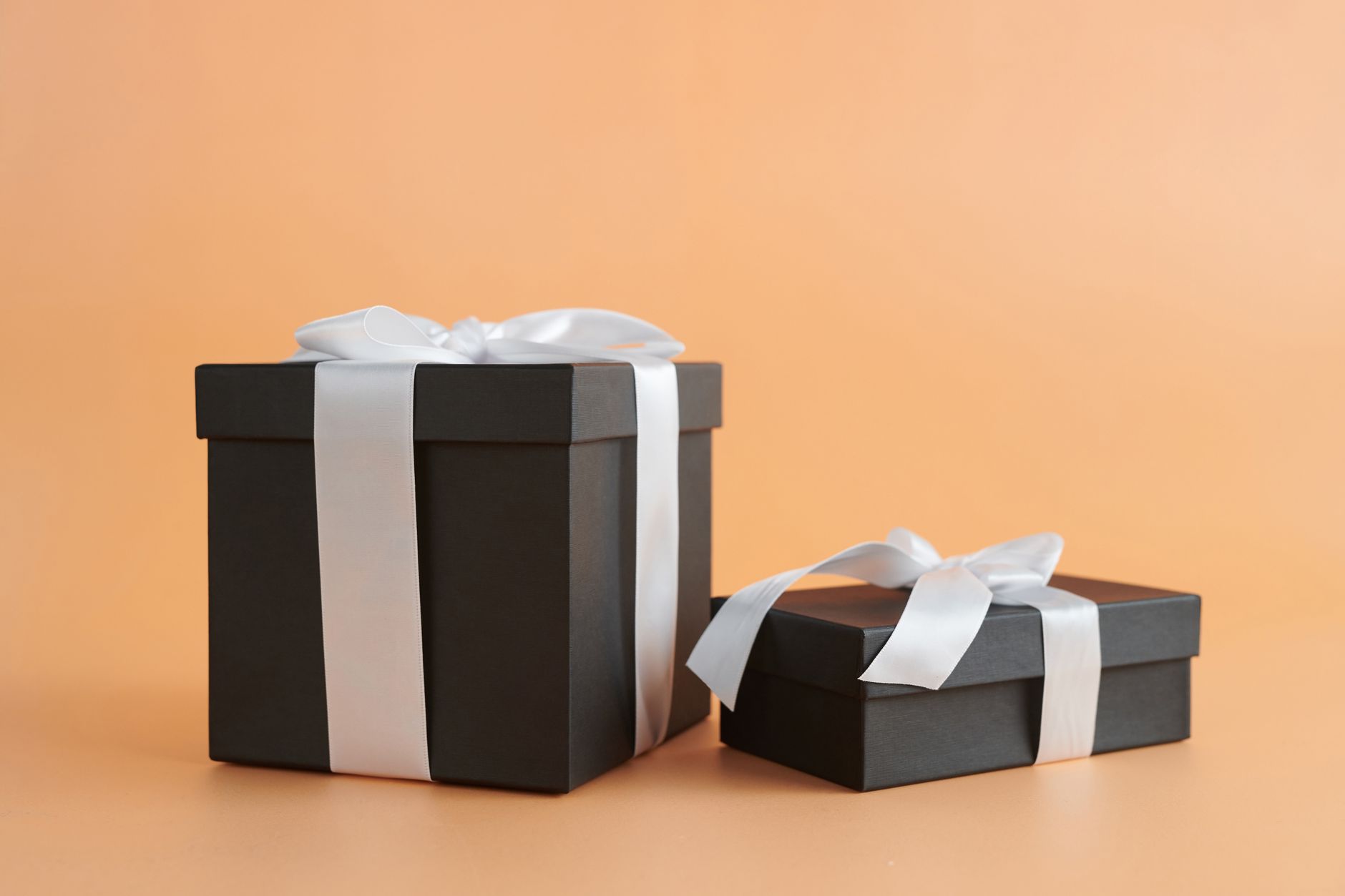 Two dark gift boxes tied with white satin ribbon on a warm peach background